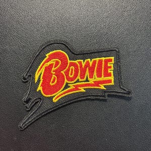 May include: An embroidered patch featuring the word Bowie in red lettering with a yellow outline. The text is set against a black background shaped like a stylized lightning bolt. The design includes a yellow lightning bolt graphic underneath the text.