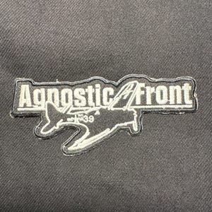 Puede incluir: Un parche negro con el texto blanco "Agnostic Front" con contorno negro. Debajo, un gráfico de avión blanco. El parche está sobre un fondo de tela negra.