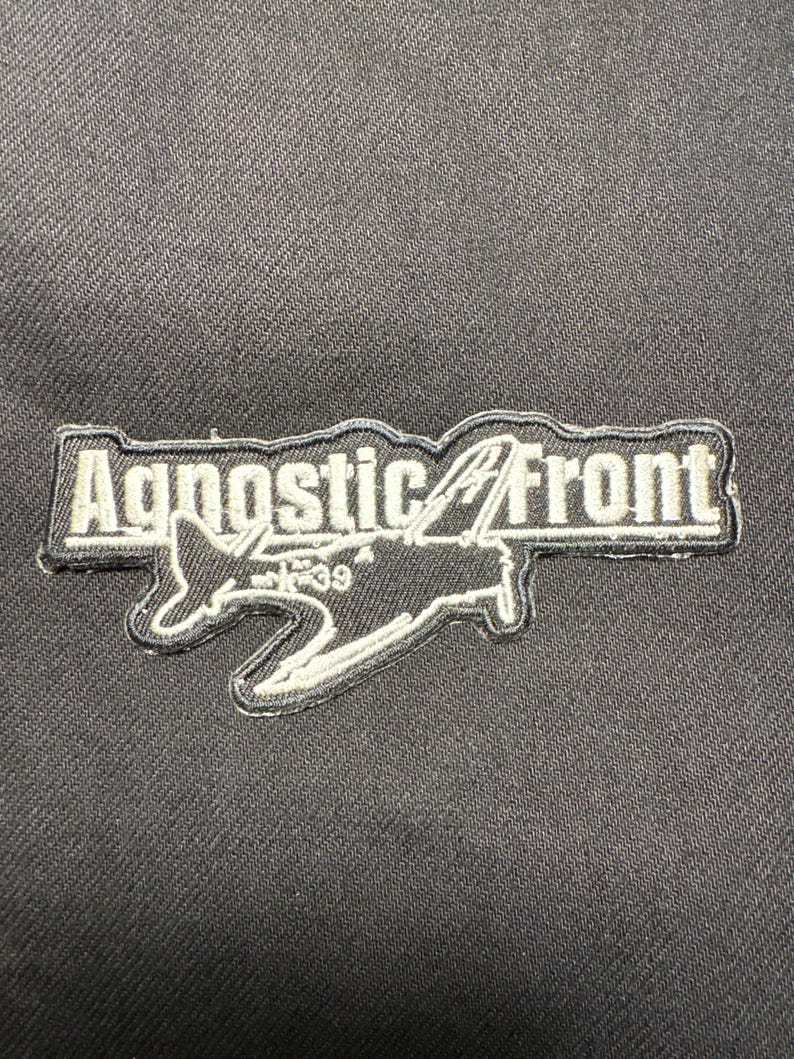 May include: A black patch with the text "Agnostic Front" in white, outlined in black. A white airplane graphic is below the text. The patch is on a black fabric background.