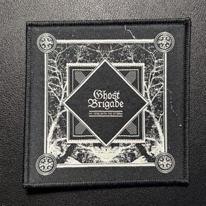 Könnte beinhalten: Ein quadratischer schwarzer Stoffaufnäher mit weißem Stickdesign, das eine zentrale Rautenform mit dem Text Ghost Brigade und IV - ONE WITH THE STORM zeigt. Der Rand enthält komplizierte Baumzweigmuster und kreisförmige Eckmotive.
