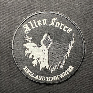 Könnte beinhalten: Ein runder, schwarzer Aufnäher mit den Worten "Alien Force" in gotischer Schrift. Der Aufnäher zeigt eine weiße, Kapuzenfigur und den Text "Hell and High Water" in Weiß.