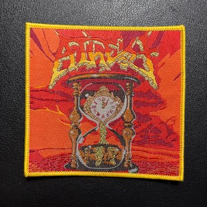 Può includere: Una toppa ricamata quadrata con bordo giallo su uno sfondo vibrante di tramonto arancione e rosso. Il design raffigura una clessidra centrale con un orologio all'interno e una scritta dorata stilizzata in alto che recita "AURA".