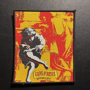 Könnte beinhalten: Quadratischer Aufnäher mit einem Guns N' Roses-Design. Der Aufnäher zeigt eine Schwarz-Weiß-Illustration einer schreibenden Person vor einem gelb-roten Hintergrund. Der Name der Band und Copyright-Informationen sind auf dem Aufnäher aufgedruckt.