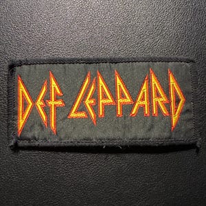 以下が含まれることがあります： 黒い長方形のパッチ。バンド名「DEF LEPPARD」が黄色とオレンジ色で刺繍されています。パッチには黒い縁取りがあり、衣服やアクセサリーに縫い付けるように設計されています。