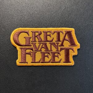 Könnte beinhalten: Ein gestickter Aufnäher mit dem Schriftzug GRETA VAN FLEET in dunkelbraunen Buchstaben. Die Buchstaben sind von einem senfgelben Rand umgeben, der der Form des Textes folgt und ein rechteckiges Emblem mit einer dicken gelben Umrandung bildet.