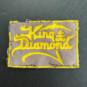 Könnte beinhalten: Rechteckiger Aufnäher mit den Worten "King Diamond" in gelben, stilisierten Buchstaben. Der Aufnäher hat einen grauen Hintergrund mit gelbem Rand. Oben auf dem Aufnäher befindet sich ein fledermausähnliches Design.