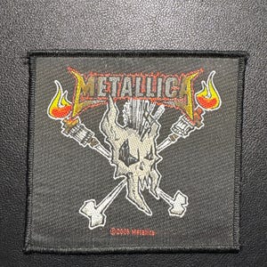 May include: Black square patch featuring the band name "Metallica" in red and yellow flame-style lettering. Below the text is a skull with crossed bones and torches. The patch is a music-themed item.
