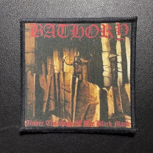 Könnte beinhalten: Ein quadratischer Stoffaufnäher mit schwarzem Stickrand, der oben den roten Schriftzug BATHORY und unten Under The Sign Of The Black Mark zeigt. Das zentrale Bild stellt eine dunkle, schattenhafte Figur in einem schwach beleuchteten Steinraum dar.