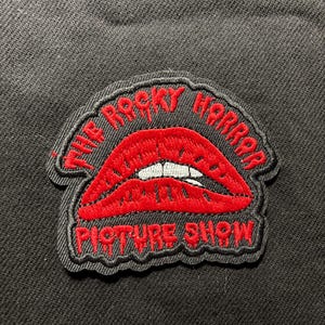 Può includere: Toppa ricamata con labbra rosse e denti bianchi, con la scritta "THE ROCKY HORROR PICTURE SHOW" in rosso. La toppa ha un bordo nero ed è posta su uno sfondo nero.