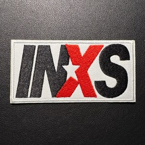 May include: Embroidered patch featuring the band name "INXS" in black and red block letters. The "X" is red with a white star in the center. The patch is rectangular with a white border, suitable for jackets, bags, or other fabric items.