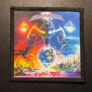 Pode incluir: Um patch quadrado com um design de fantasia colorido. O patch apresenta uma imagem central de um rosto monstruoso, um globo e o texto "No World Order!". A parte superior do patch tem a palavra "Gamma Ray" em uma fonte estilizada.
