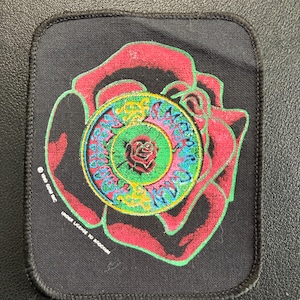 Könnte beinhalten: Ein rechteckiger schwarzer Stoffaufnäher mit einer großen roten Rosengrafik und grünem Umriss. In der Mitte befindet sich ein kreisförmiges Emblem mit einer kleineren Rose, umgeben von gelbem und blauem Text, der Grateful Dead lautet. Enthält 1990 GDAS Inc.