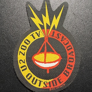 Könnte beinhalten: Schwarzer Aufnäher mit gelbem und rotem Text und Grafiken. Der Text lautet "U2 ZOO TV OUTSIDE BROADCAST". Ein gelber Blitz befindet sich über einem rot-gelben Satellitenschüssel-Design. Der Aufnäher ist von 1992.