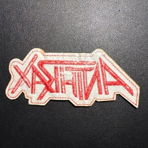 May include: A white patch with the word "ANTHRAX" in red, stylized lettering. The patch has a red stitched border and is likely designed to be sewn onto clothing or accessories. The patch is on a dark surface.