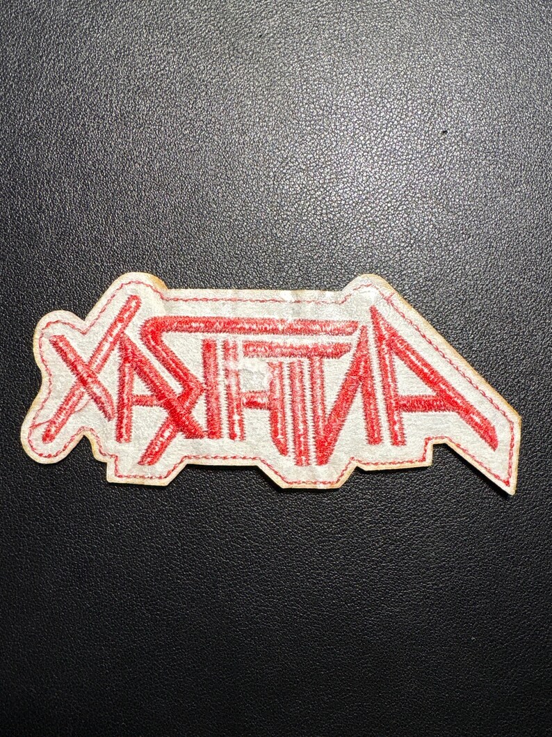 May include: A white patch with the word "ANTHRAX" in red, stylized lettering. The patch has a red stitched border and is likely designed to be sewn onto clothing or accessories. The patch is on a dark surface.