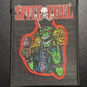 Op de afbeelding: Zwarte rechthoekige patch met de rode woorden "SPITE FUEL" en een schedel erboven. De patch toont een groen monster met geel haar, dat een microfoon vasthoudt en een rock and roll handgebaar maakt.