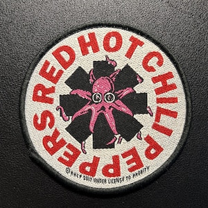 Può includere: Una toppa ricamata circolare con un simbolo nero a forma di asterisco su sfondo bianco. Un polpo rosa è centrato sopra il simbolo. Il testo RED HOT CHILI PEPPERS è ricamato in rosso lungo il bordo, con il testo del copyright in basso.