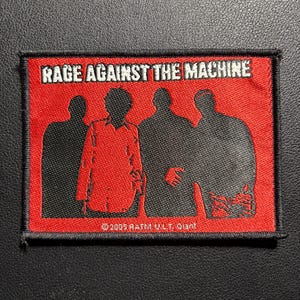 Könnte beinhalten: Ein rechteckiger Aufnäher mit dem Text "RAGE AGAINST THE MACHINE" in Weiß auf rotem Hintergrund. Darunter befinden sich vier schwarze Silhouetten von Personen. Der untere Teil des Aufnähers trägt den Text "©2005 RATM V.L.T. Giant."