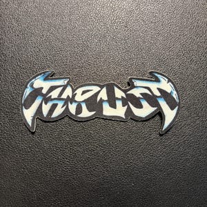 May include: A fabric patch featuring the word THRUST in a stylized, graffiti-inspired font. The lettering is primarily white with black outlines and a light blue gradient effect, creating a sharp, angular appearance with pointed ends.