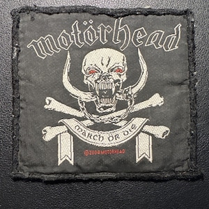 Könnte beinhalten: Schwarzer quadratischer Aufnäher mit dem Bandnamen "Motörhead" in weißen gotischen Buchstaben über einem Totenkopf mit Hörnern, gekreuzten Knochen und den Worten "March or Die". Der Aufnäher ist mit schwarzen Nähten eingefasst und enthält einen Copyright-Hinweis.