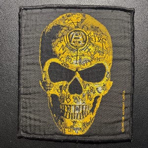 May include: A rectangular black fabric patch featuring an embroidered yellow skull design. The skull is covered in intricate occult-style symbols and text. The text 2003 THE ALCHEMY CARTA is embroidered in small yellow letters on the lower right side.