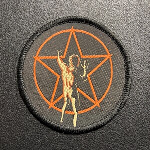 May include: Circular black patch featuring an orange pentagram design. Within the pentagram is an orange and tan illustration of a person with raised arms. The patch has a black border, set against a black background.