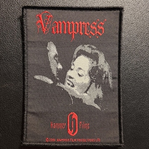 May include: A rectangular black embroidered patch featuring the red text Vampress at the top. Below is a grayscale graphic of two figures. The bottom includes the red text Hammer Films with a logo and the text 2008 HAMMER FILM PRODUCTIONS LTD.