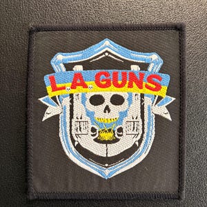 Könnte beinhalten: Schwarzer quadratischer Aufnäher mit einem Totenkopf-Design in einem Schild. Der Text "LA GUNS" ist in Rot und Gelb gehalten. Der Aufnäher hat einen blauen und weißen Rand.