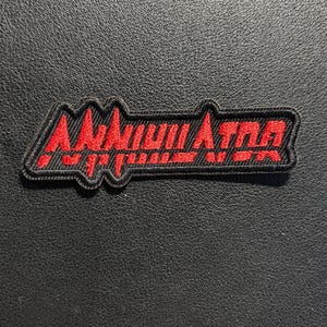 May include: A rectangular embroidered patch with a black background and a red border. The patch features the word ANNIHILATOR stitched in bold red capital letters with a stylized, jagged font. The design has a distinct, sharp, and angular aesthetic.