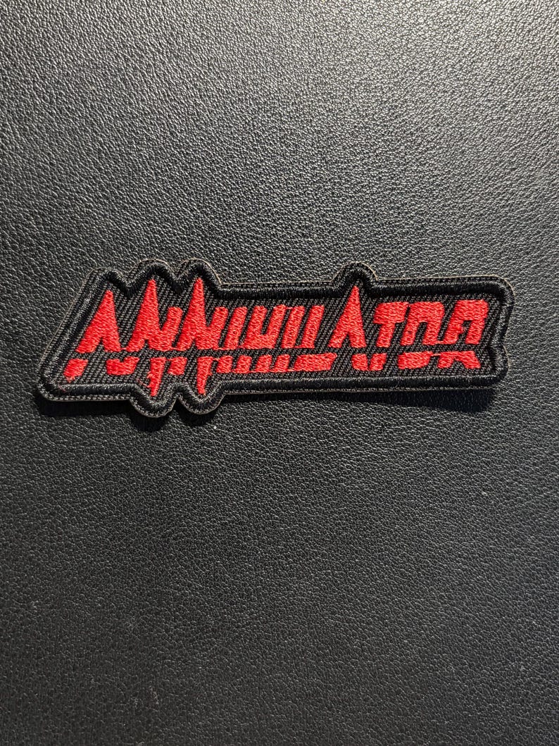 May include: A rectangular embroidered patch with a black background and a red border. The patch features the word ANNIHILATOR stitched in bold red capital letters with a stylized, jagged font. The design has a distinct, sharp, and angular aesthetic.