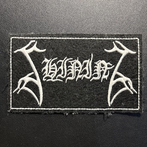 May include: A rectangular black fabric patch featuring white embroidered text that reads SHINING in a stylized gothic font. The design includes symmetrical white embroidered decorative elements on the left and right sides, all enclosed within a white stitched border.