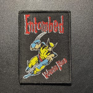 May include: A rectangular embroidered patch with a black background. The top features the word Entombed in red text with white outlines. Below is a graphic of the character Wolverine in yellow and blue, and the text Wolverine Blues in red.