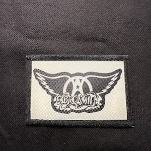 May include: Black fabric with a rectangular patch featuring the Aerosmith logo. The patch has a white background with a black winged design and the band's name in a stylized font. The patch is bordered by a black trim.