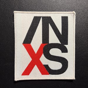 May include: Square white patch with the letters ANXS in black and red. The letters AN are black, the X is red, and the S is black. The patch has a white border and is on a black surface.