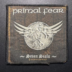 Puede incluir: Un parche rectangular con las palabras "primal fear" en blanco, fuente estilizada. Debajo, un emblema plateado con alas y el texto "Seven Seals". El parche tiene un borde negro y un fondo marrón.
