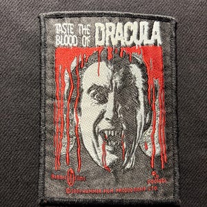 May include: Embroidered patch with a portrait of Dracula, red blood drips, and the text "TASTE THE BLOOD OF DRACULA." The patch has a black border and is set against a dark background. The design is a vintage horror movie theme.