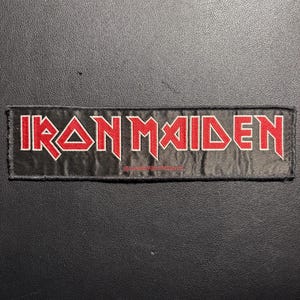 Könnte beinhalten: Ein rechteckiger schwarzer Aufnäher mit dem roten Schriftzug "IRON MAIDEN". Der Text ist in einer fetten, stilisierten Schriftart gehalten. Der Aufnäher ist wahrscheinlich aus Stoff und hat einen genähten Rand. Dies ist ein Band-Merchandise-Artikel.