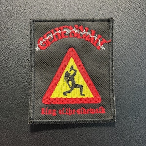 Op de afbeelding: Een rechthoekige zwarte geborduurde patch met een rood en geel driehoekig waarschuwingsbord-icoon met een zwart silhouet van een drinkend persoon. De tekst "GENEWALK" staat in het rood erboven en "king of the sidewalk" in het rood eronder.