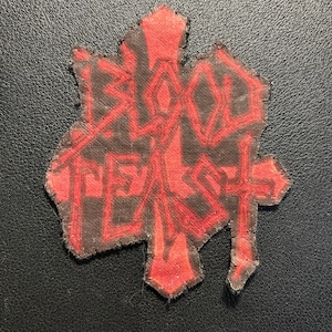 Könnte beinhalten: Ein Stoffaufnäher mit schwarzem Hintergrund und rotem gesticktem Text, der BLOOD FEAST lautet. Der Aufnäher hat eine unregelmäßige, gezackte Form mit einem kreuzartigen Design, das in den Schriftzug integriert ist, sowie eine Textur.