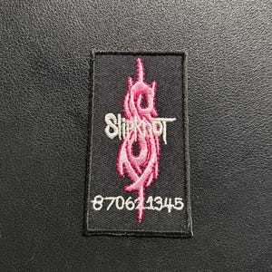 May include: A rectangular black embroidered patch featuring the pink Slipknot logo in the centre, the word Slipknot in white text to the left, and the white numerical sequence 870621345 embroidered at the bottom of the design.