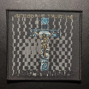 Puede incluir: Parche negro y gris con un fondo de patrón de cuadros. Una cruz detallada con una figura está centrada, con detalles en azul y dorado. El texto "IN GOD WE TRUST, INC." está en la parte superior y "PAUL KING INC." en la parte inferior.