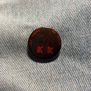 Könnte beinhalten: Ein runder Ansteckbutton mit schwarzem Hintergrund und roter Schrift, die "LET THERE BE ROCK" und "AC/DC" in der Mitte zeigt. Das Design enthält eine schwache rote grafische Umrisslinie eines Gitarristen über dem Bandnamen.