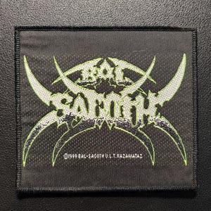 Peut inclure: Un écusson carré en tissu noir avec un logo brodé en fil vert clair et blanc. Le motif présente le texte BAL-SAGOTH dans une police stylisée et pointue avec des accents incurvés. Sous le logo, le texte indique 1999 BAL-SAGOTH U.L.T.RAZAMATAZ.
