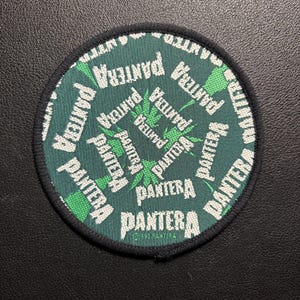 Könnte beinhalten: Runder Aufnäher mit dem Wort "PANTERA" in Weiß auf dunkelgrünem Hintergrund, kreisförmig angeordnet. Grüne Cannabisblatt-Designs sind ebenfalls vorhanden. Der Aufnäher hat einen schwarzen Rand.