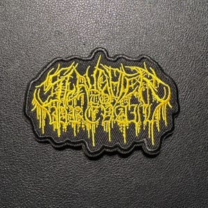 May include: A black embroidered patch with yellow text. The text is stylized and appears to be a band name or logo. The patch has a black border and is set against a textured black background.