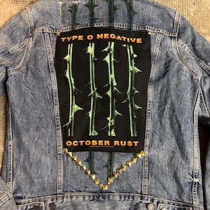 Könnte beinhalten: Blaue Jeansjacke mit einem großen schwarzen Aufnäher auf dem Rücken mit dem Text "TYPE O NEGATIVE OCTOBER RUST" in Orange. Der Aufnäher hat ein grünes und schwarzes Dornen-Design. Goldfarbene Nieten sind in Dreiecksform angeordnet.