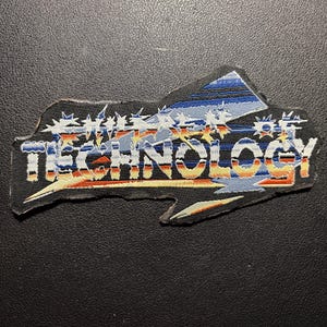 Può includere: Una toppa nera con la scritta "TECHNOLOGY" in bianco, blu e arancione. La toppa ha un design a bordo frastagliato con una forma a montagna blu e arancione nella parte superiore. Lo sfondo è nero.