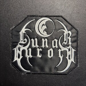 May include: Black and white embroidered patch with the text "Lunar Aurore" in a gothic font. The patch has a unique octagonal shape and features a crescent moon design. The patch is likely for clothing or accessories.