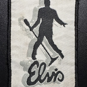 May include: Embroidered patch featuring a black silhouette of Elvis Presley performing with a microphone. The image is set against a light gray background with a shadow effect and the name "Elvis" in a stylized script.