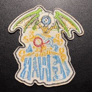 K&ouml;nnte beinhalten: Gestickter Aufn&auml;her mit einem detaillierten Design mit einem zentralen Augenmotiv, flankiert von gr&uuml;nen und gelben Fl&uuml;geln. Der Aufn&auml;her enth&auml;lt blaue, gelbe und rote Akzente mit blauem Text am unteren Rand. Der Aufn&auml;her hat einen wei&szlig;en Rand.